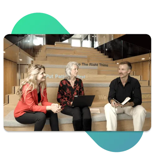 Two women and one man sitting on wooden steps in a modern office, engaged in discussion with a laptop and notebook.