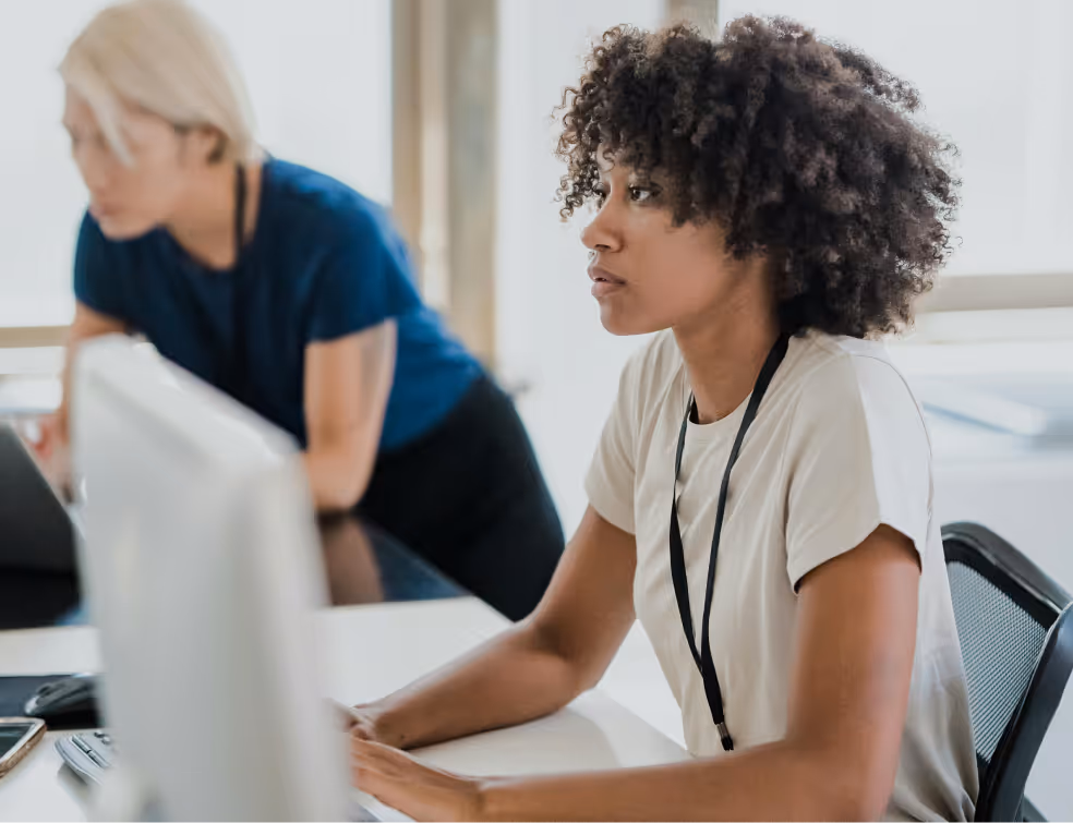 **Alt text:**
Two professionals working at a desk in an office. The person in the foreground is focused on a computer screen, while the person in the background leans forward, also engaged in work.